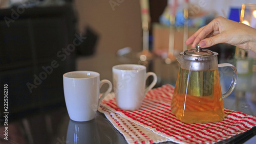 A hand closes the lid of the kettle. Glass teapot beside white mugs, tea preparation and loose leaf brewing in hospitality and food and beverage service.