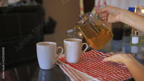 A hand pours tea—steeped from a glass teapot—into white ceramic mugs, cups meant for a cozy home atmosphere, hospitality, and the serving of food and drinks at home.
