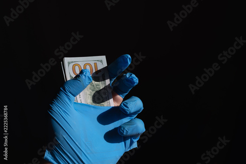 Hand in a torn medical glove holding a 100 dollar bill on a black background.