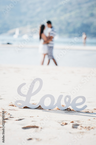 Fotografie Romantic couple embraces on a sunlit beach behind a white Love sign in the sand, evoking summer romance, intimacy, travel and dreamy seaside wedding or engagement moments