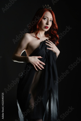 Red haired woman in black sheer fabric with tiara, dramatic studio lighting against dark background