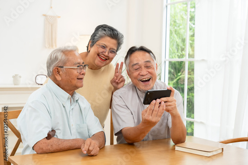 Three senior Asian residents smile while using smartphone for video call in assisted living center, enjoying digital communication, companionship, friendship, social engagement, joyful active aging.