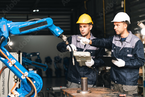 Industrial engineer and technician inspect robotic arm in factory workshop, discussing automated manufacturing equipment and safety procedures during collaborative maintenance work