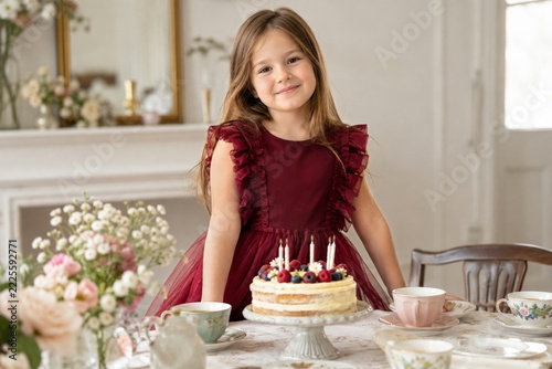 Girl in burgundy party dress behind birthday cake and colorful teacups
