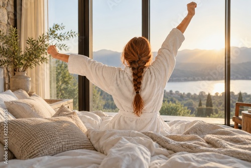 Woman in a white robe stretches toward sunlit windows above soft bedding