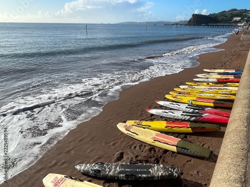 Colorful rescue and racing surfboards line a sandy beach beside rolling waves on a bright coastal day