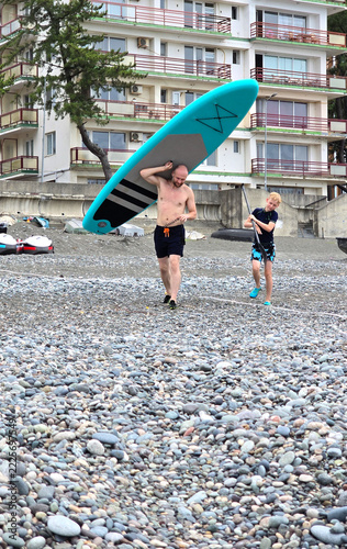 A father and son are walking along the beach, carrying a SUP board.