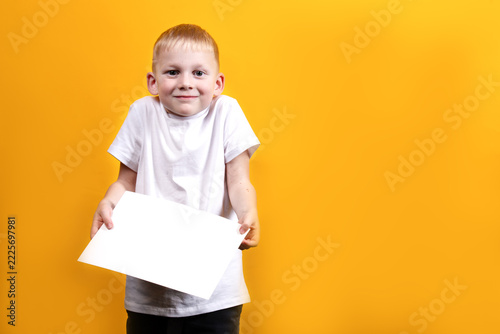 A boy in a white T-shirt holds a white sheet of paper against a yellow background and looks surprised.