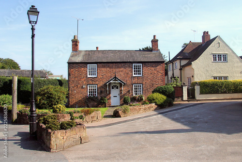 Traditional English village street with a historic brick cottage, period homes, landscaped gardens and a heritage-style street lamp on a sunny day.