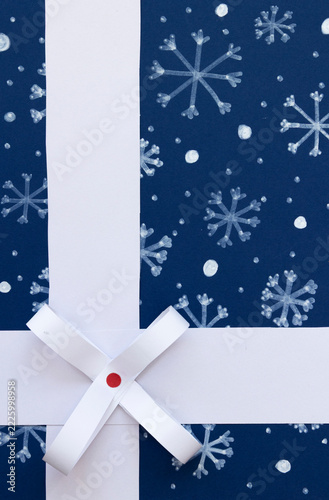 Paper Christmas gift with a white ribbon and paper bow on a dark blue background with drawn snowflakes. Visual resource with copy space, ideal for advertising and commercial design