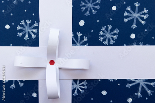 Paper gift with a white ribbon and decorative bow on a dark blue background with drawn snowflakes and copy space. Christmas concept for advertising and commercial design
