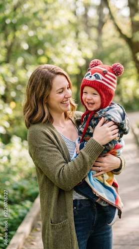 Happy Mother Holding Adorable Baby Boy in Cozy Hat Outdoors in Green Nature