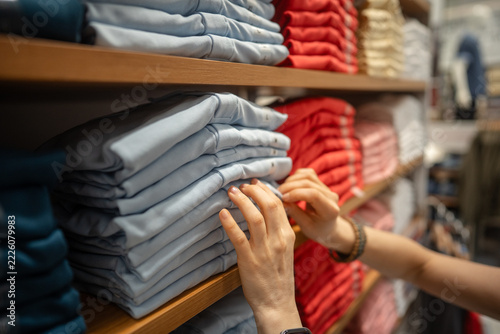 Woman purchasing new basic clothing at garment fashion showroom, woman looking through stack of t-shirts on shelf display, examinees quality of material, fabric and preferable size of clothing item.