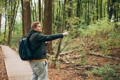 Blonde young 30s man wearing backpack holding phone and pointing something on city autumn park street. Urban lifestyle, travel concept. High quality photo