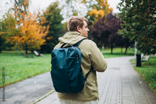 A man with backpack walking on the street in the city park. Urban lifestyle concept. Traveler. Autumn time. Young male in casual clothing standing outdoor. Rear view
