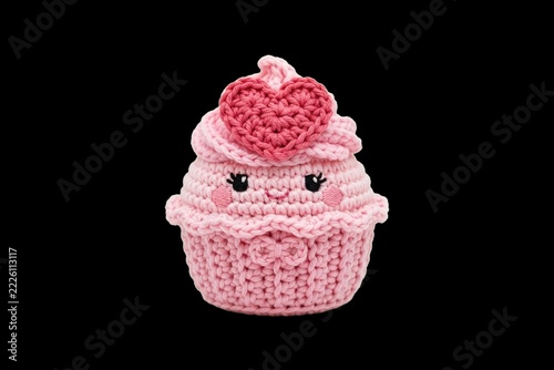 Handmade amigurumi crochet art of a smiling pink cupcake character with a heart on its frosting, isolated on black for Valentine's Day crafts and sweet celebrations.