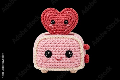 Handmade amigurumi crochet character of a smiling pink toaster popping out a red heart-shaped toast against a black background for a Valentine's Day romance and affection concept.