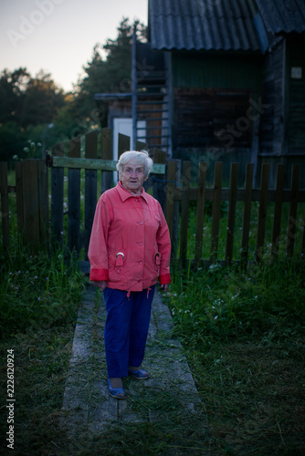 Elderly woman standing on a narrow wooden path beside a rustic house and fence in evening light
