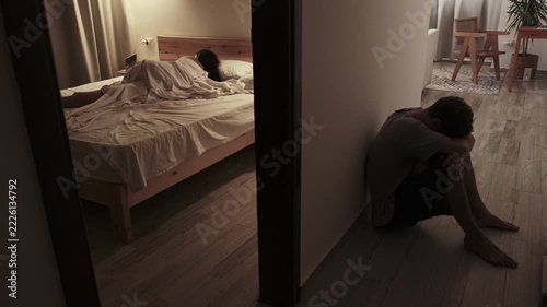 Emotionally withdrawn man sitting on the floor with head down while his partner lies turned away in bed, portraying relationship crisis, silent treatment and emotional pain at home