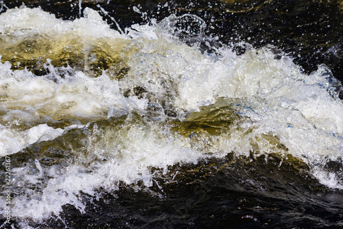 Turbulent river water crashing over rocky terrain creates powerful white foam and dynamic liquid splashes in this close-up view of a wild natural stream during sunny summer day