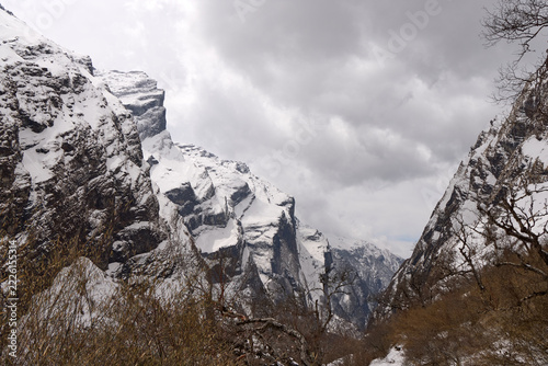 A trekking route winds through a valley in the Himalayas. Snow covers the ground and mountains, leading toward Annapurna Base Camp in Nepal under a cloudy sky