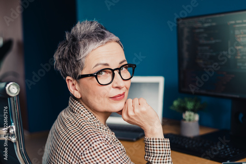 Mature woman programmer wearing glasses looks over shoulder in home office with laptop and monitor displaying code, confident professional at desk