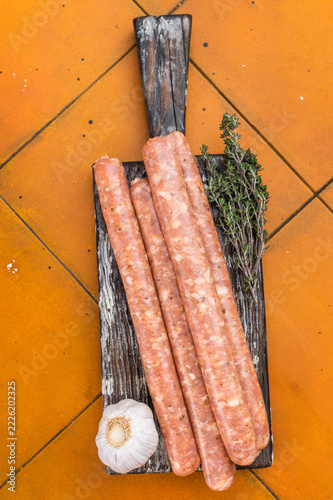 Fotografie Fresh uncooked Merguez sausages with herbs on wooden board over orange tiles