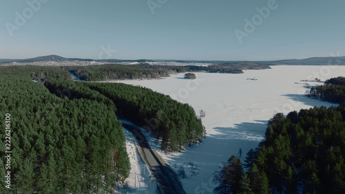 An elevated winter view follows a curved route through evergreen woodland beside an icebound lake, while snowfields and clear daylight reveal the remote landscape of the Ural Mountains, Russia