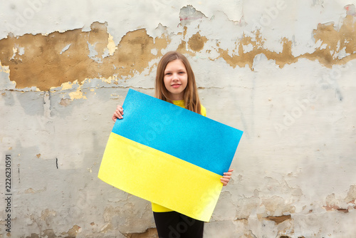 Young girl holds a large poster of the Ukrainian flag on a weathered wall