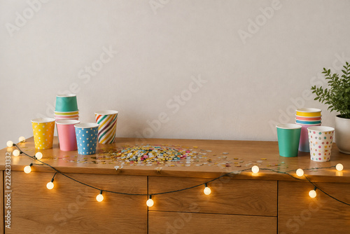 Wooden sideboard with paper cups, metallic confetti and string lights, party decoration concept