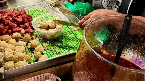 Close-up of a street food vendor serving assorted cooked meatballs and pieces of meat from a display tray into a plastic takeaway container at a local food market in Thailand. Traditional Asian street