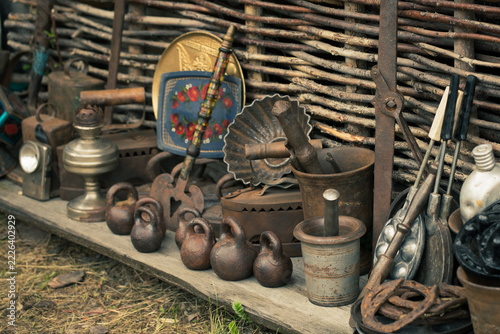 Vintage forged metal tools and antique household items displayed at a local blacksmith craft fair against a rustic wattle fence, selective focus.