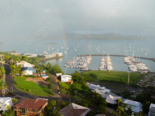 Aerial view of Airlie Marina with yachts docked in the calm harbor, surrounded by lush green hills, residential houses, and a faint rainbow in the sky, Airlie Beach, Queensland, Australia.