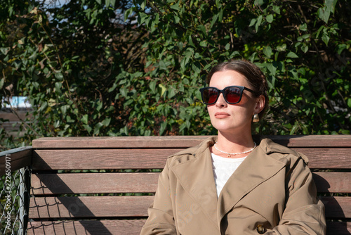 Photography Woman in trench coat sits on wooden bench wearing sunglasses beside leafy shrubs