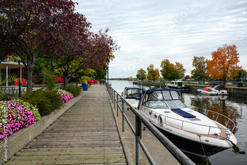 Wooden boardwalk follows Sainte-Anne-de-Bellevue Canal as pedestrians walk past moored boats and flower beds beneath textured clouds in muted daylight.
