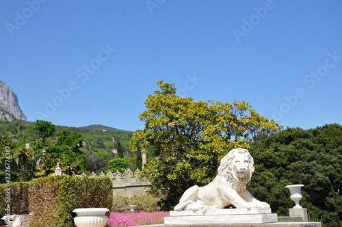 Majestic marble lion statue in lush palace garden, surrounded by trees, flowers and mountains, symbol of strength in a sunny landscape