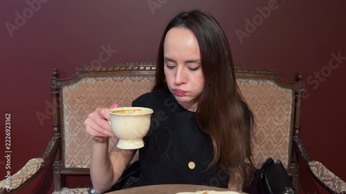Young woman in black top sits in vintage chair drinking from white cup with eyes closed. Dark red background, cafe interior, coffee break, relaxation, enjoying drink