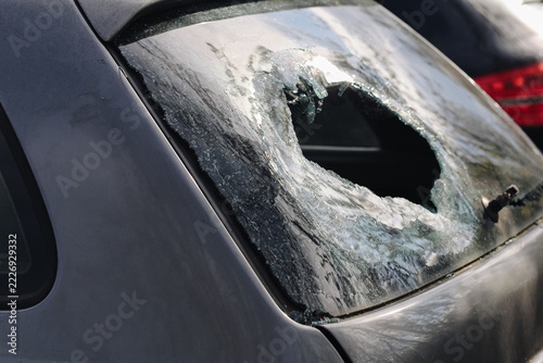 Broken rear car window. Large hole in shattered glass. Severe vehicle window damage. Jagged fragments around central opening. Automotive vandalism and repair concept. Cracked windshield background.