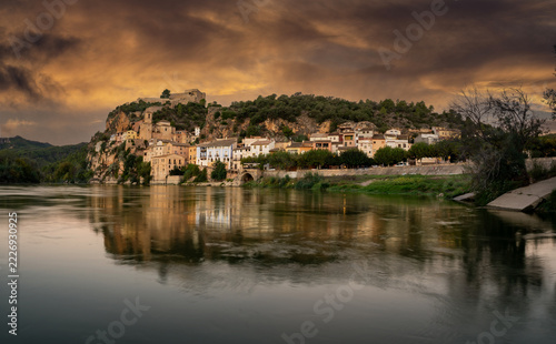 A picturesque historic village with an ancient fortress nestled atop a steep rocky cliff, perfectly reflected in the calm waters of a wide river under a bright blue sky  
