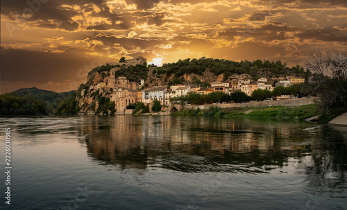 A picturesque historic village with an ancient fortress nestled atop a steep rocky cliff, perfectly reflected in the calm waters of a wide river under a bright blue sky  