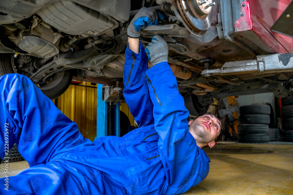 Obraz premium Caucasian male mechanic in blue mechanic jumpsuit lying flat on his back while inspecting the undercarriage of a raised car in a busy auto repair shop