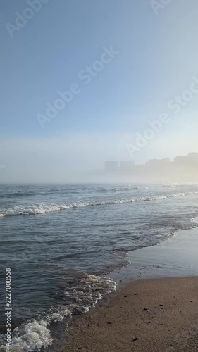 Atmospheric seaside cityscape in sea fog, empty sand beach with rolling waves