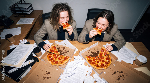 Late Night Grind: Two exhausted colleagues find solace and sustenance in a late-night pizza feast amidst a chaotic office environment, fueled by caffeine and shared desperation.