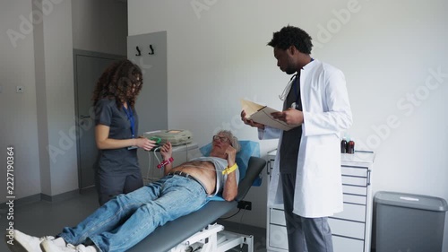 A medical team prepares a senior man for an ECG examination in a clinic. A female healthcare worker attaches wrist electrodes while a male doctor reviews a medical folder.