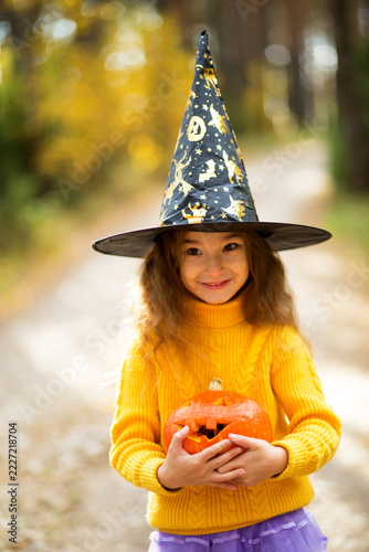 A cheerful young girl wearing a pointy witch hat and holding a festive orange pumpkin outdoors in a sunny autumn park.