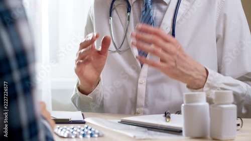 A doctor in a white lab coat with a stethoscope talks to a patient at the office desk in a clinic. In the foreground are a blister pack of pills, white medicine bottles, and a medical notepad.