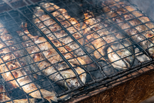 Obraz na plátně Detailed macro shot of whole fresh fish being carefully cooked in a traditional metal grill basket over an open charcoal fire with thick smoke highlighting rustic culinary techniques