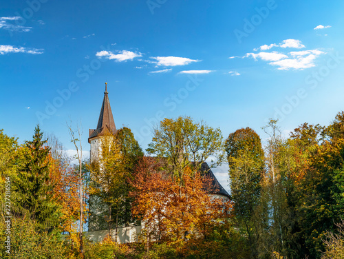 Historic Church Tower Peeking Through Autumn Trees Under A Bright Blue Sky In Rural Landscape