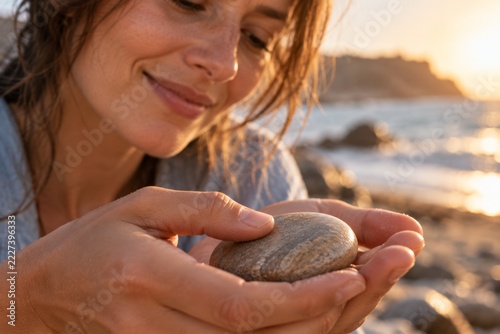 A woman cradles a smooth beach stone in her palm and traces its sun warmed surface beside the ocean. Tactile mindfulness inspired by simple natural details