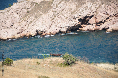 Scenic coastal view with traditional wooden boat on blue sea near rocky cliffs and dry summer grassland, natural landscape under sunny weather